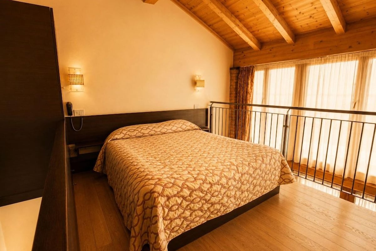 Une chambre dotée d’un grand lit, de poutres apparentes en bois au plafond, d’un balcon avec une balustrade et de deux appliques murales.