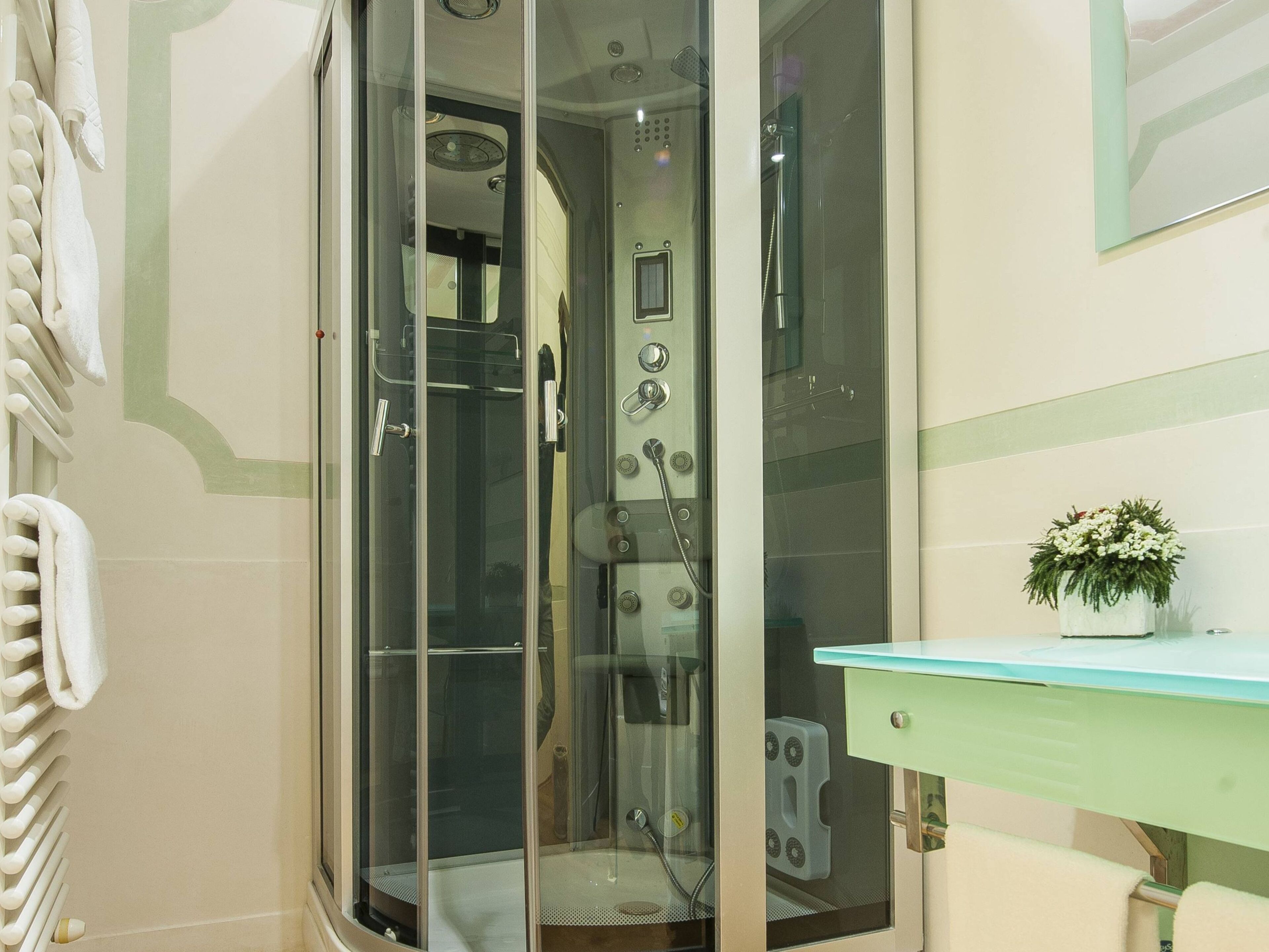A glass-enclosed shower with a control panel, a towel rack, and a small potted plant on a light-colored vanity.