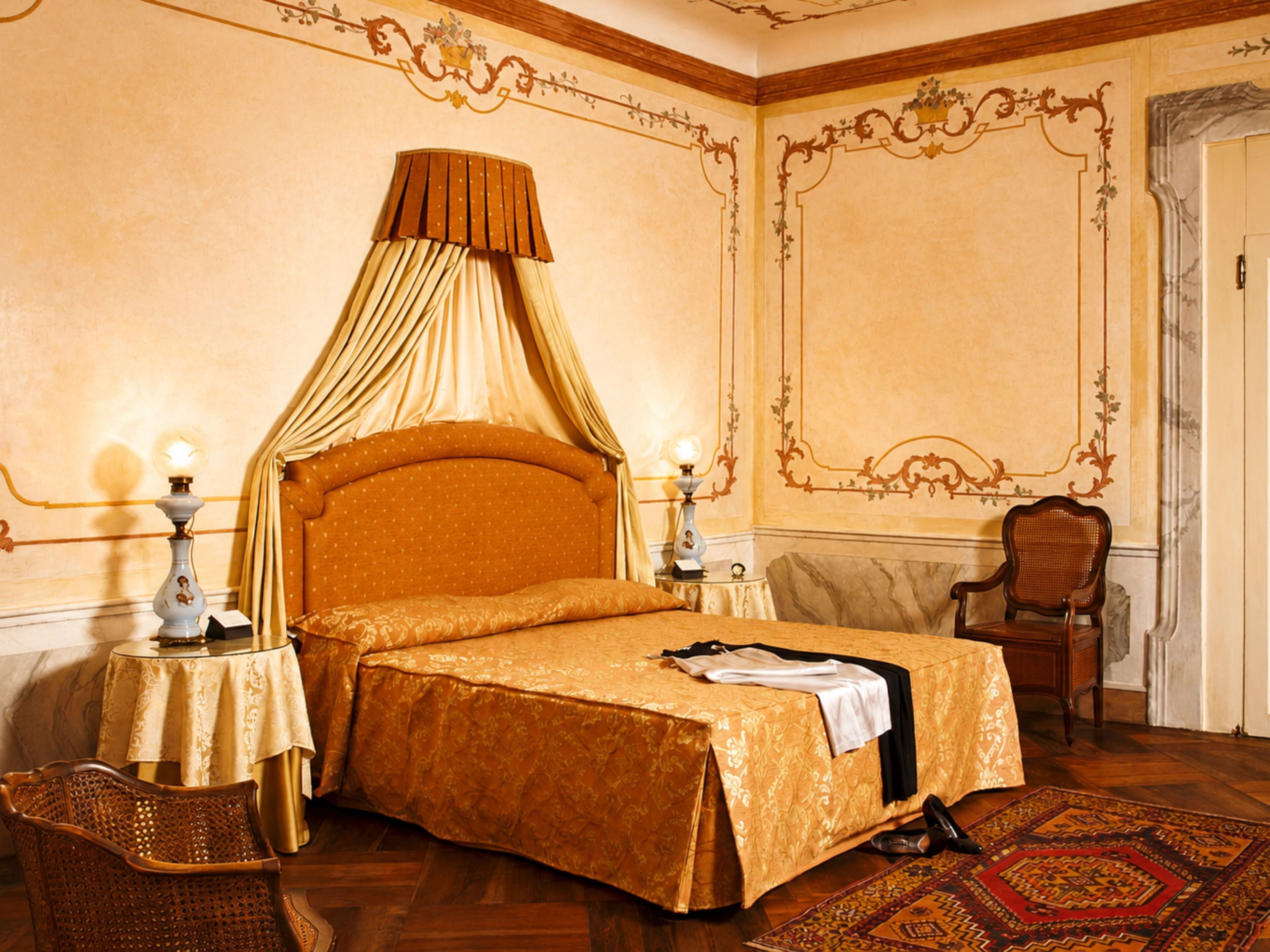 Ein Zimmer mit einem Himmelbett, zwei Nachttischen, einem Sessel und einem gemusterten Teppich.