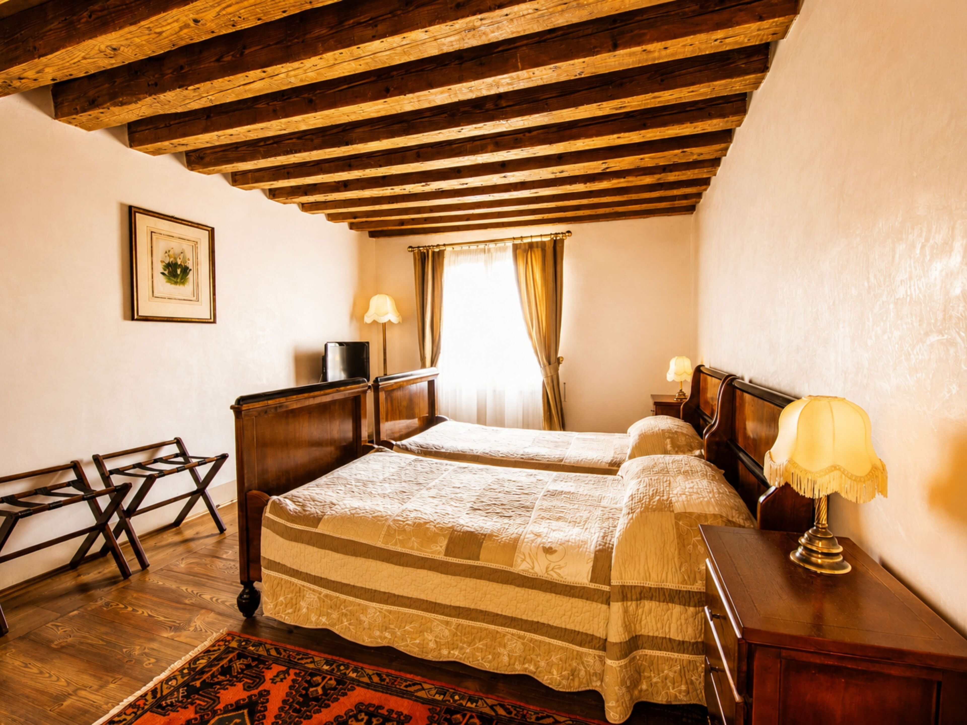 A room with two beds, wooden beams on the ceiling, a desk, a chair, a lamp, and a window with curtains.