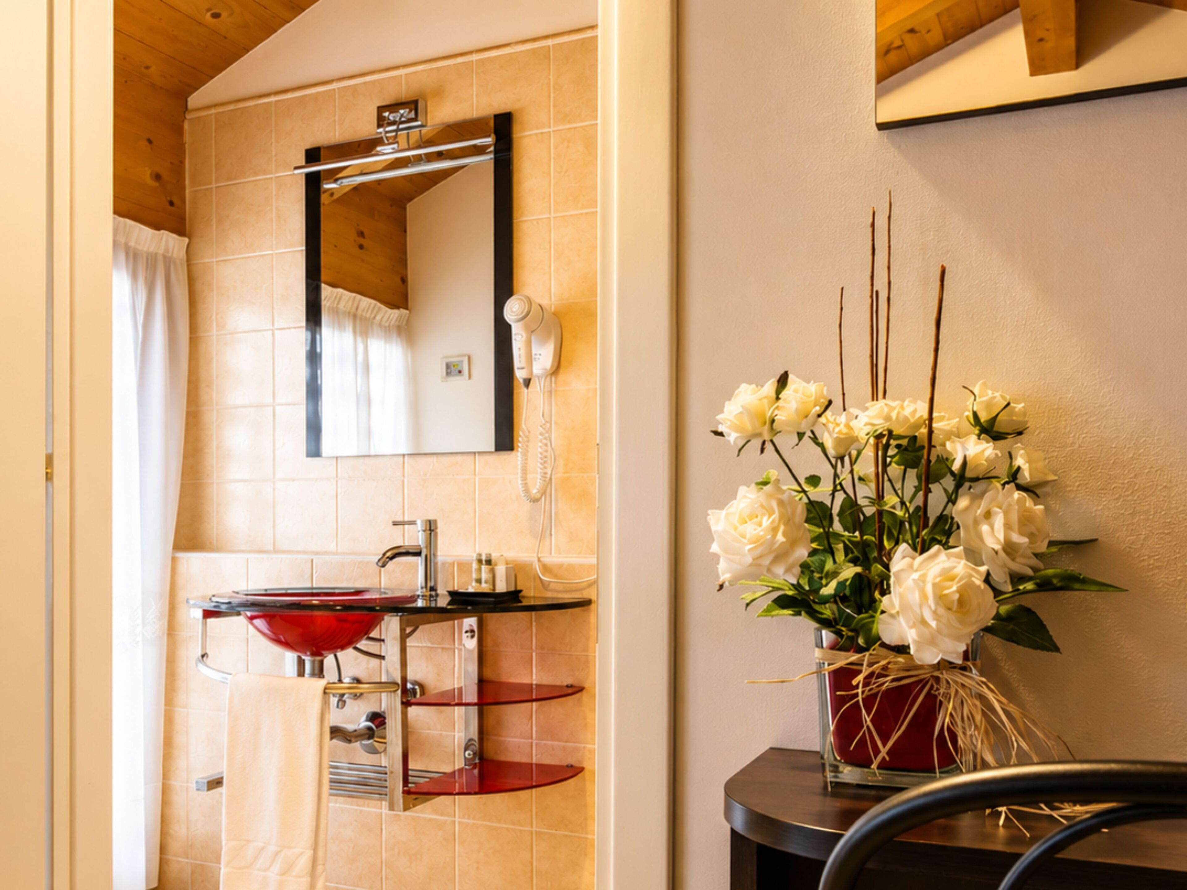 A bathroom with a red sink, a mirror, a hairdryer, and a vase of white flowers.