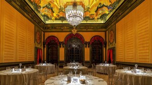 A grand ballroom with ornate ceiling, chandeliers, and round tables set for dining.