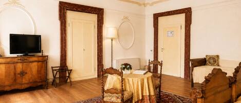 Une pièce avec un plancher en bois, un lit avec une tête de lit, un bureau avec une lampe, un téléviseur posé sur une commode en bois et un grand miroir accroché au mur.