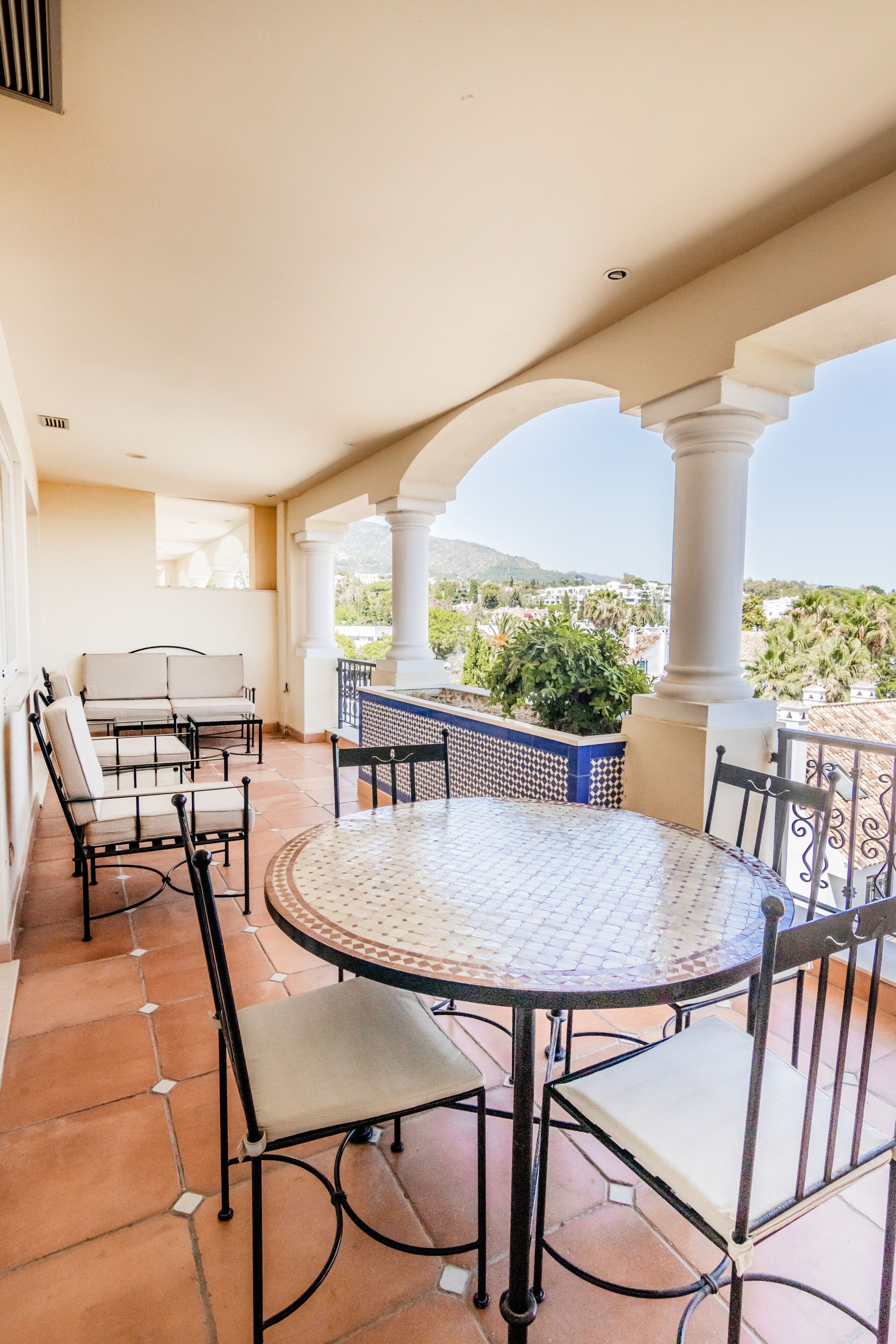 Un patio cubierto con una mesa redonda y sillas, vistas a zonas verdes y edificios a lo lejos, y suelo de baldosas.