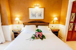 Una habitación de hotel con una cama grande, dos mesitas de noche con lámparas, una obra de arte enmarcada sobre la cama y una puerta de madera con un panel de vidrio.