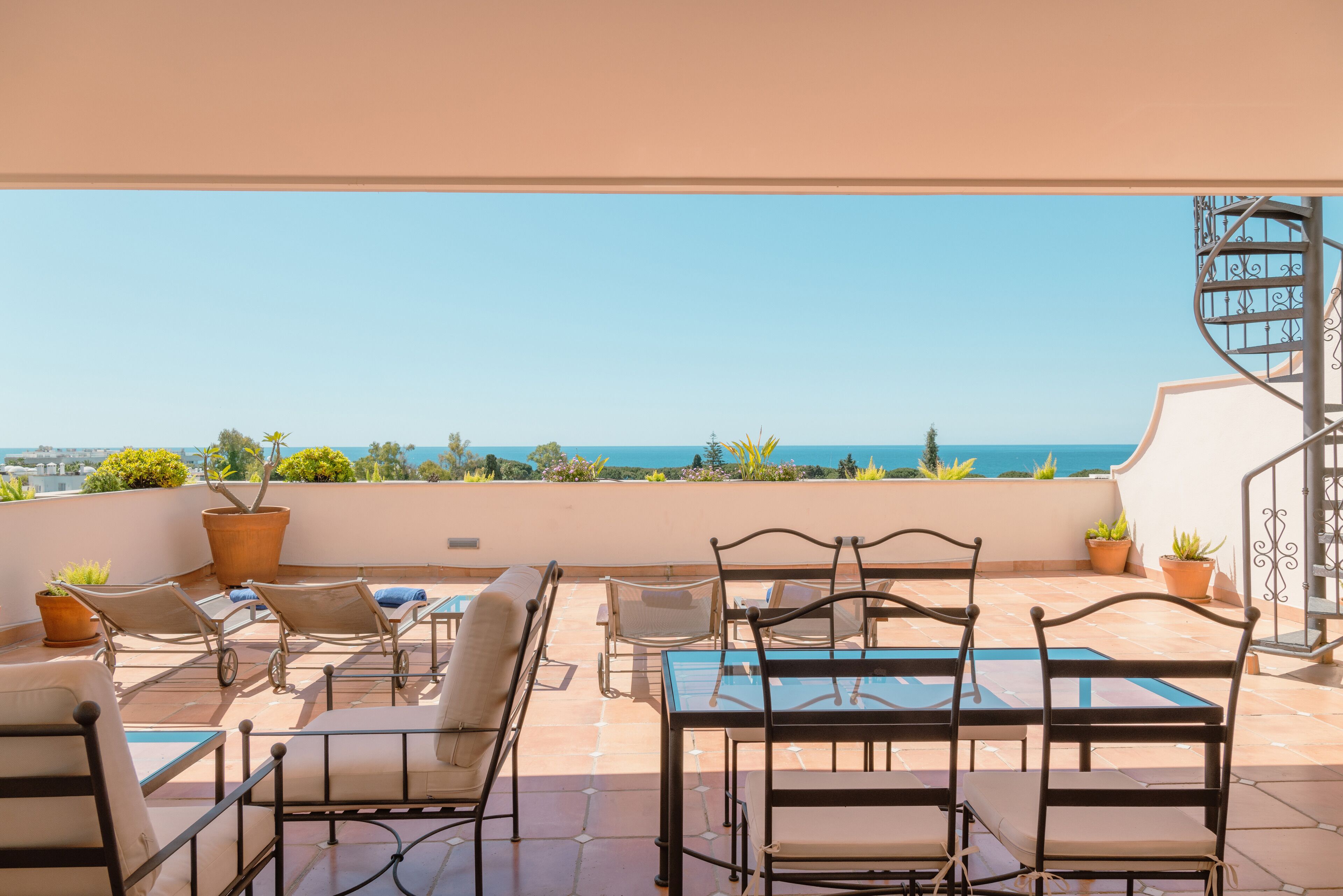 Une terrasse avec une table et des chaises en verre, des plantes en pot et une vue sur la mer.