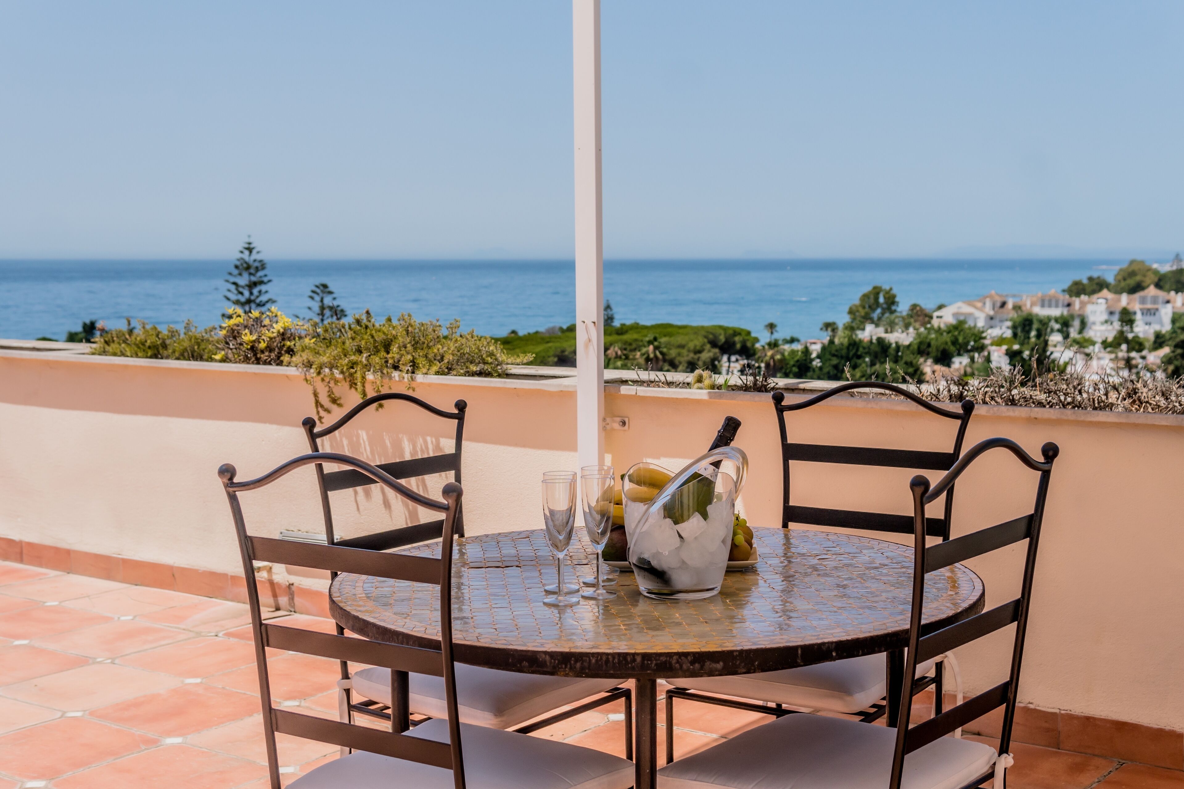Un tavolo rotondo con quattro sedie, una bottiglia di champagne e una ciotola di frutta su una terrazza con vista sul mare.