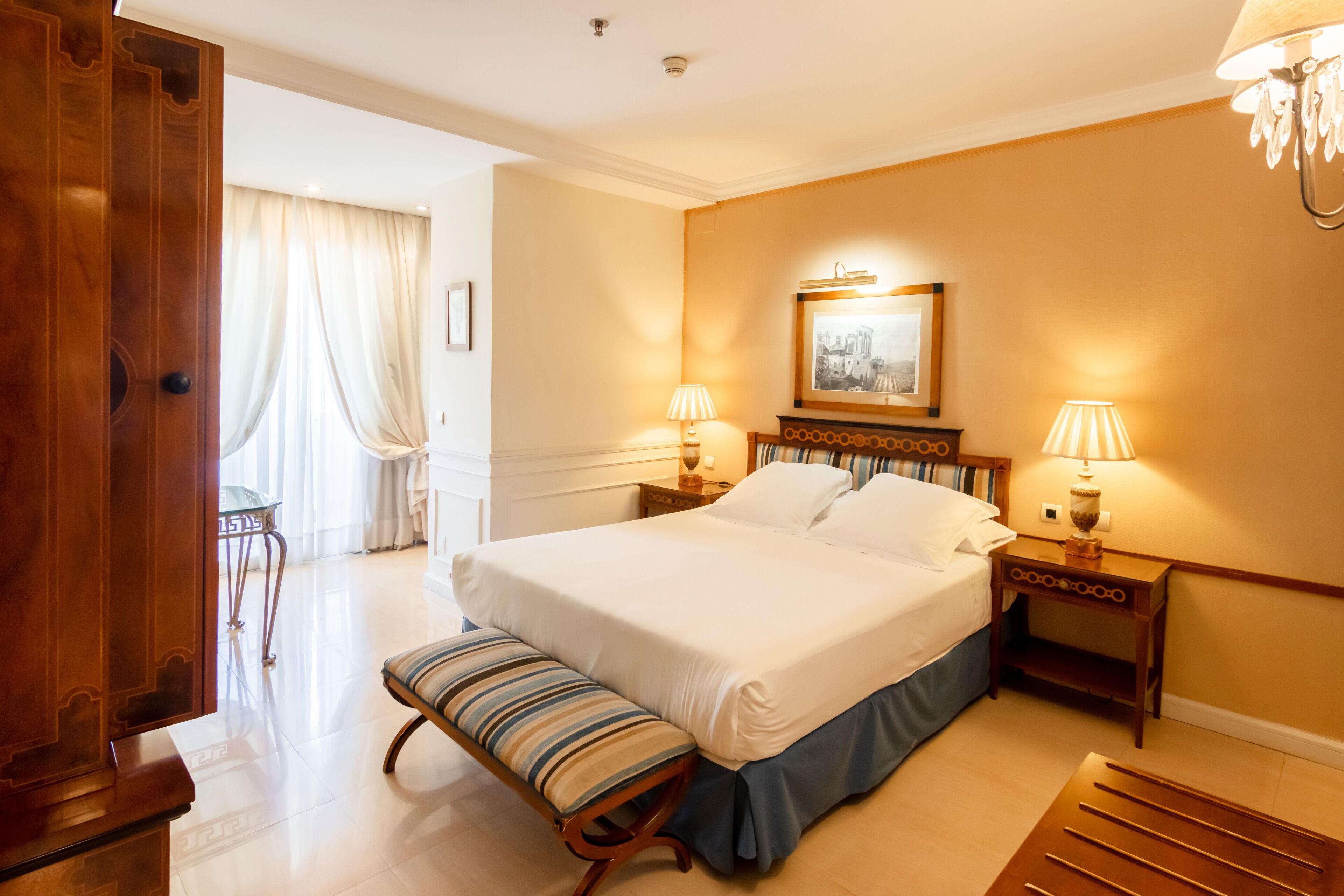 Een hotelkamer met een groot bed, twee nachtkastjes met lampen, een bankje, een houten garderobe en uitzicht buiten door een raam met gordijnen.