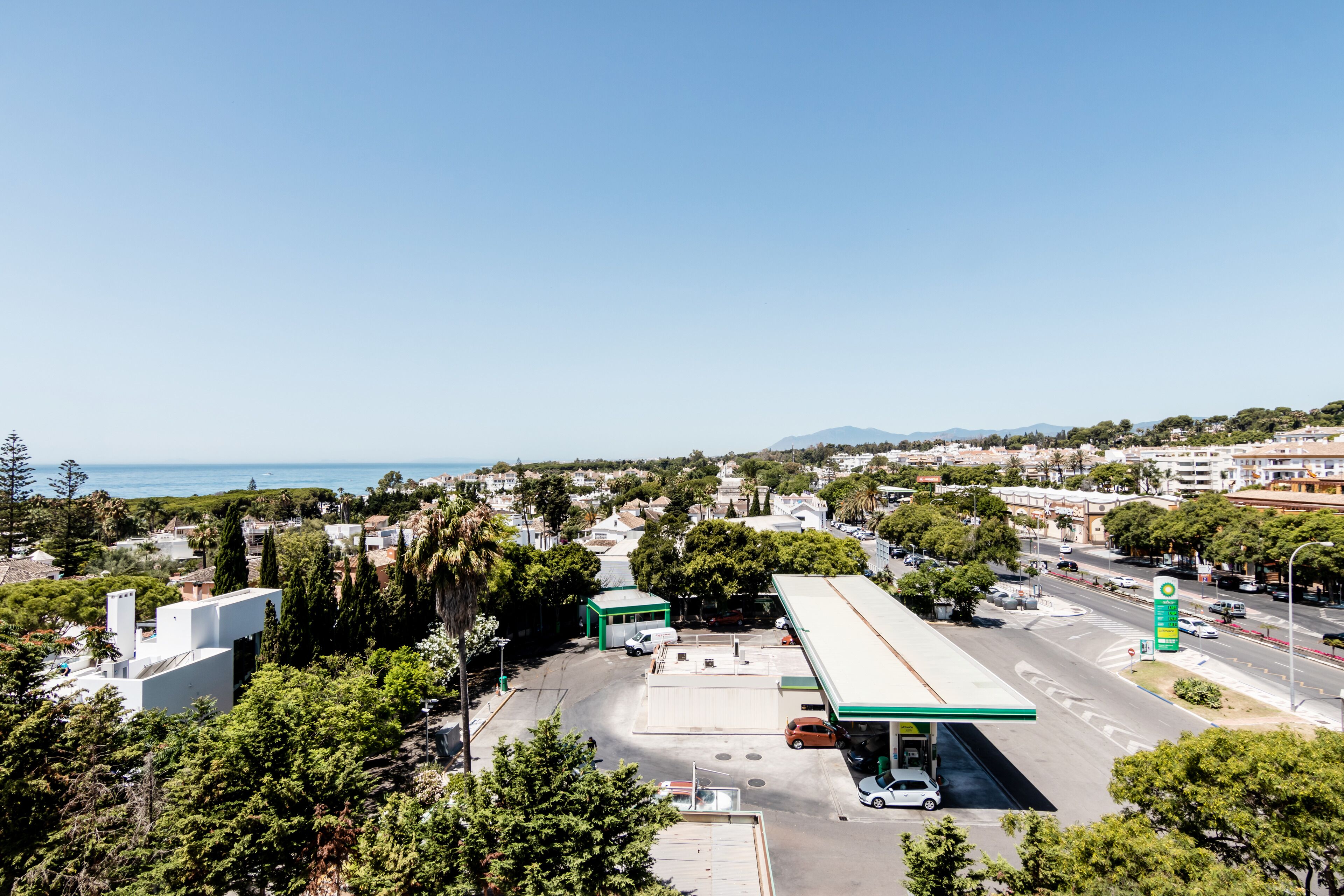 Una estación de servicio con un toldo verde, rodeada de árboles y edificios, y una vista despejada del mar al fondo.