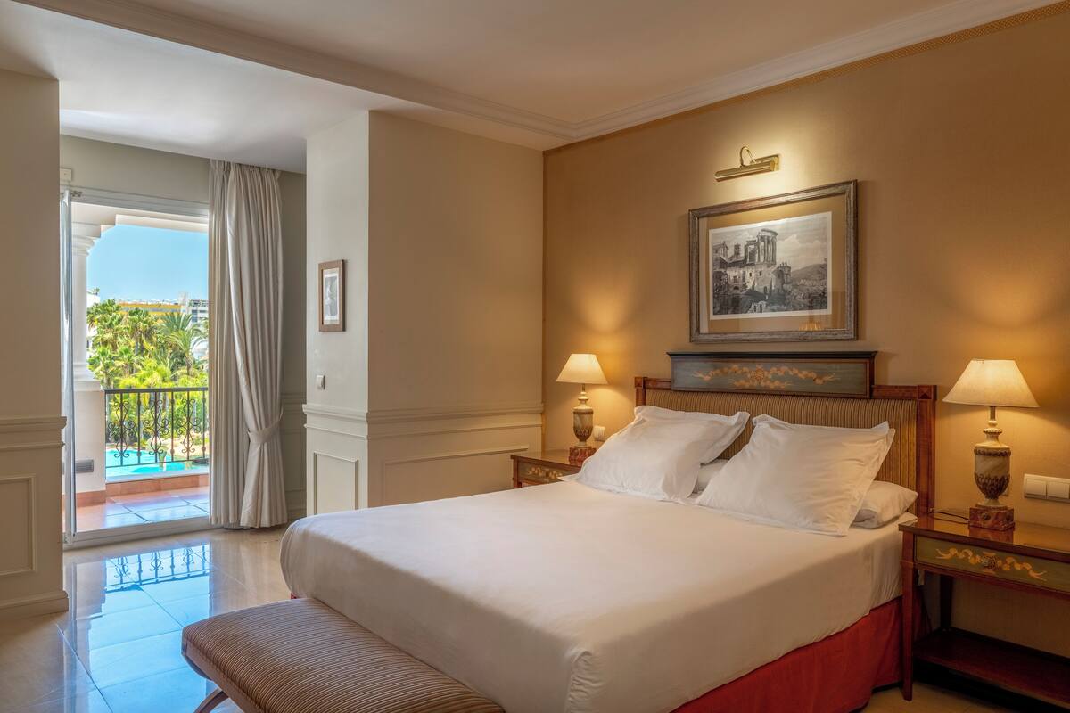 A hotel room with a large bed, two bedside lamps, a framed picture on the wall, and a balcony with curtains.