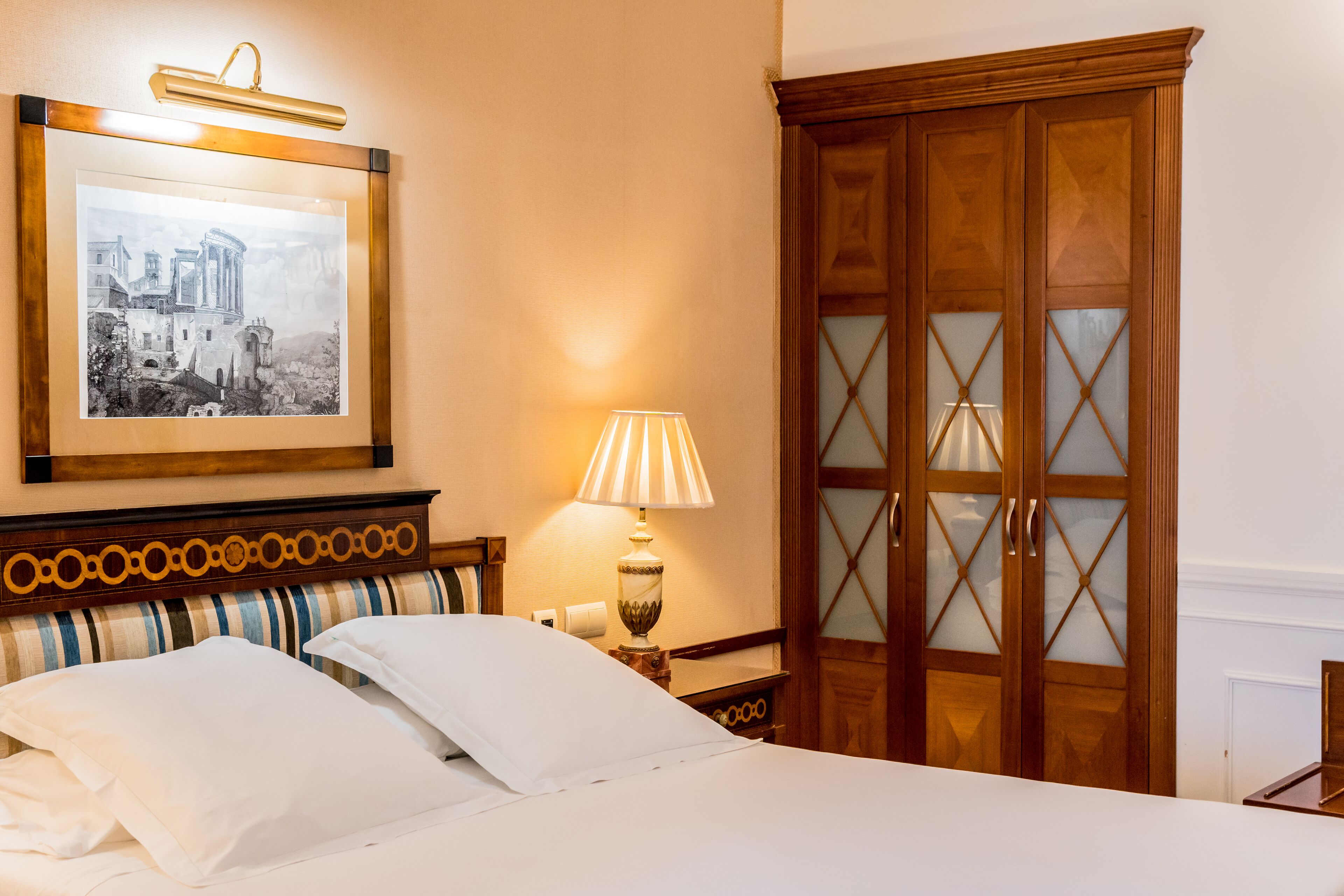 Una cama bien hecha con ropa de cama blanca, una fotografía en blanco y negro de un paisaje urbano enmarcada sobre ella, un armario de madera con paneles de vidrio y una lámpara de noche.