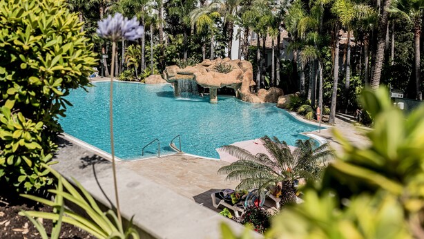 A pool area with a waterfall feature, surrounded by palm trees and a rocky landscape. - Guadalpin Suites (Marbella)