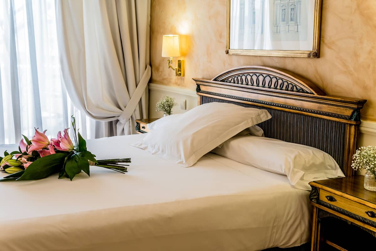 A neatly made bed with white linens and a bouquet of pink flowers.