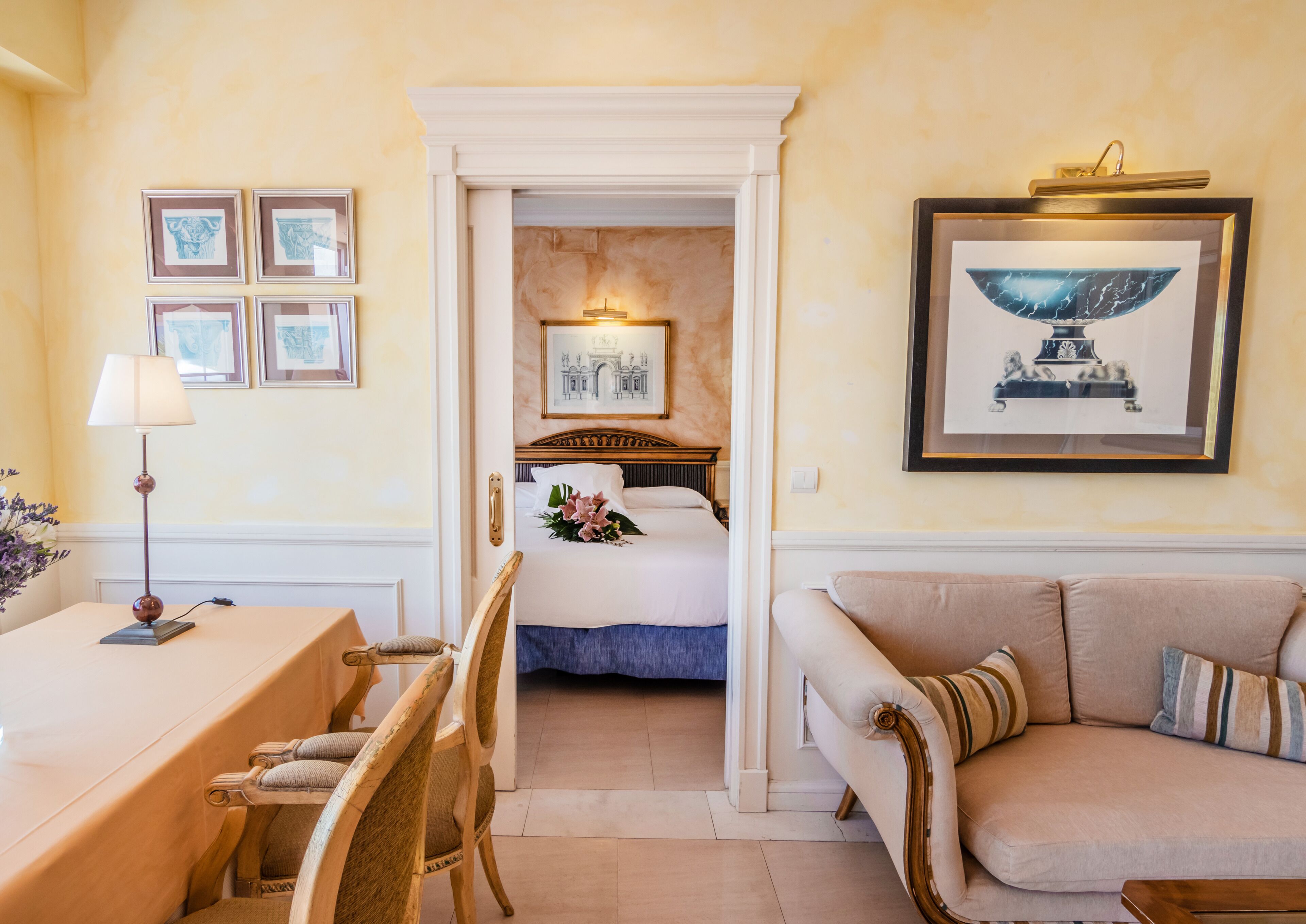 A hotel room with a dining area, a sofa, a bed with a floral arrangement, and framed artwork on the walls.