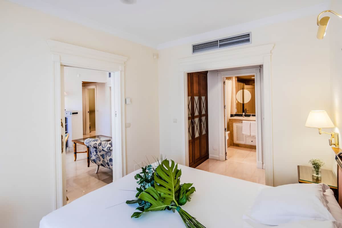 A hotel room with a bed, a nightstand with a lamp, a table with a chair, and a bathroom visible through an open door.
