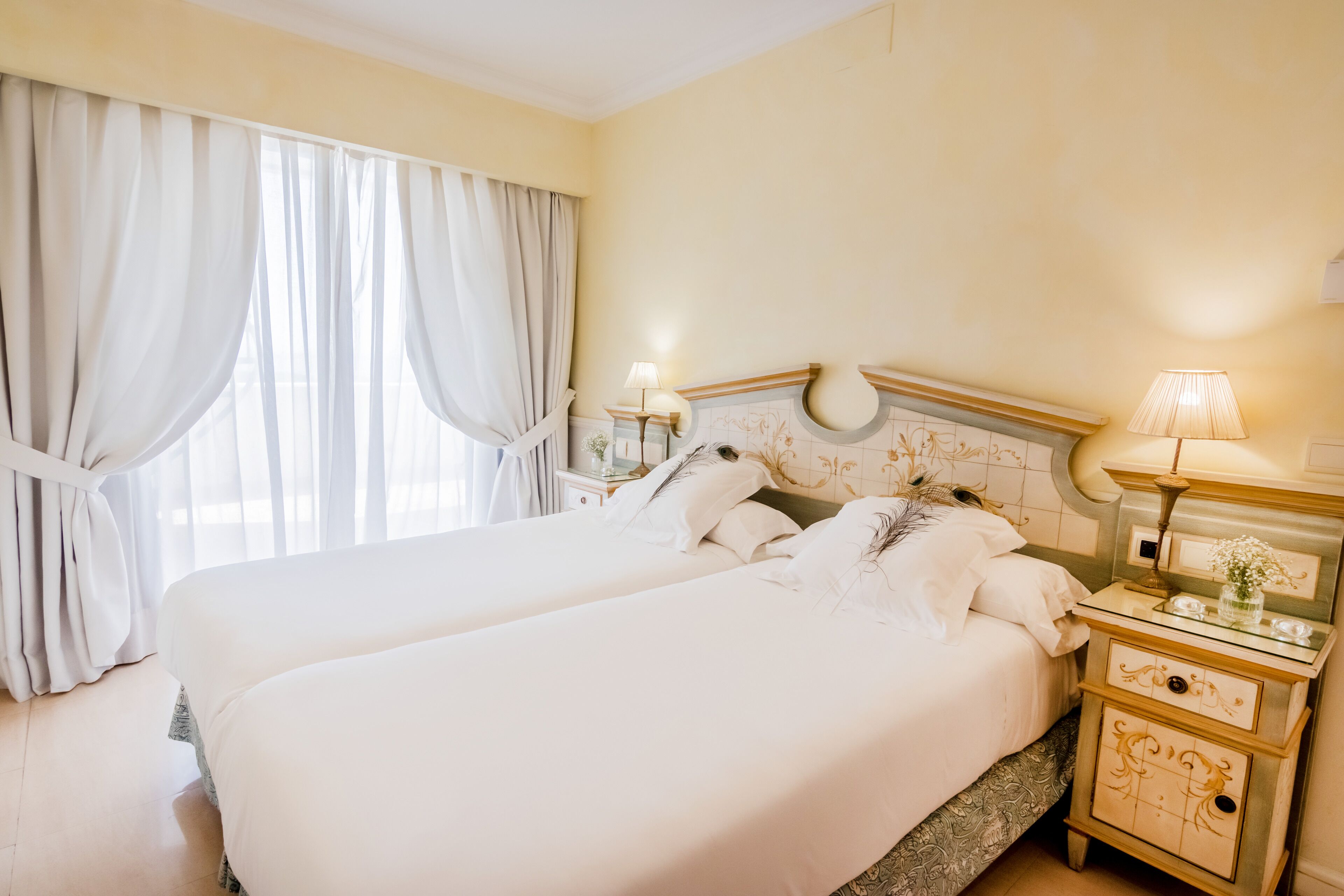 A double bed with white linens, a nightstand with a lamp, and a window with sheer curtains.