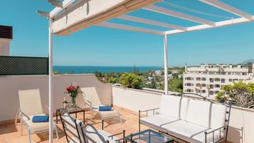 A terrace with a white sofa, two armchairs, and a glass table, overlooking a cityscape and the sea.