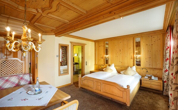 Une chambre aux murs lambrissés, avec un grand lit, un lustre et un bureau en bois recouvert d’une nappe.