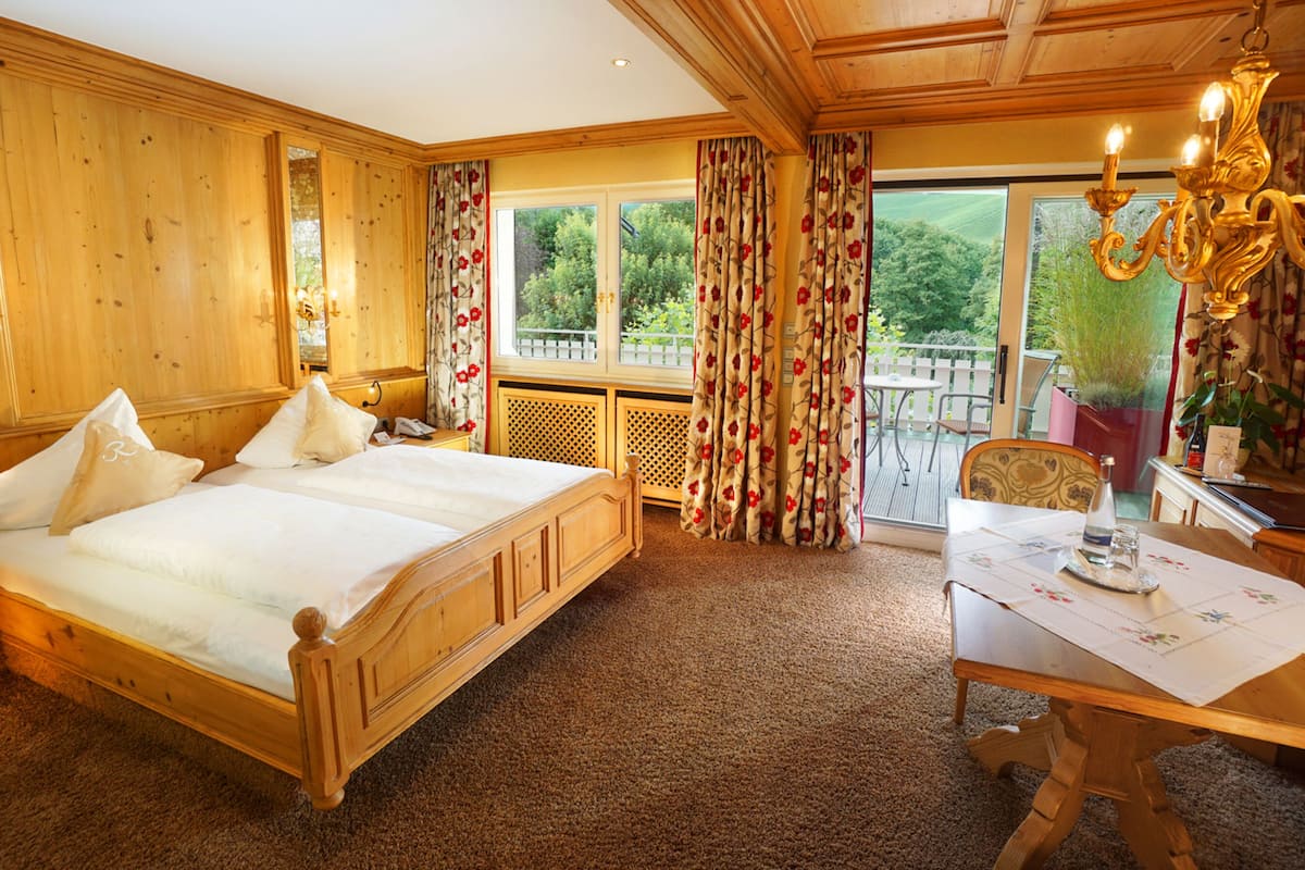 Ein Schlafzimmer aus Holz mit einem großen Bett, einem Schreibtisch mit Stuhl und einem Balkon mit Blick auf Grünflächen.