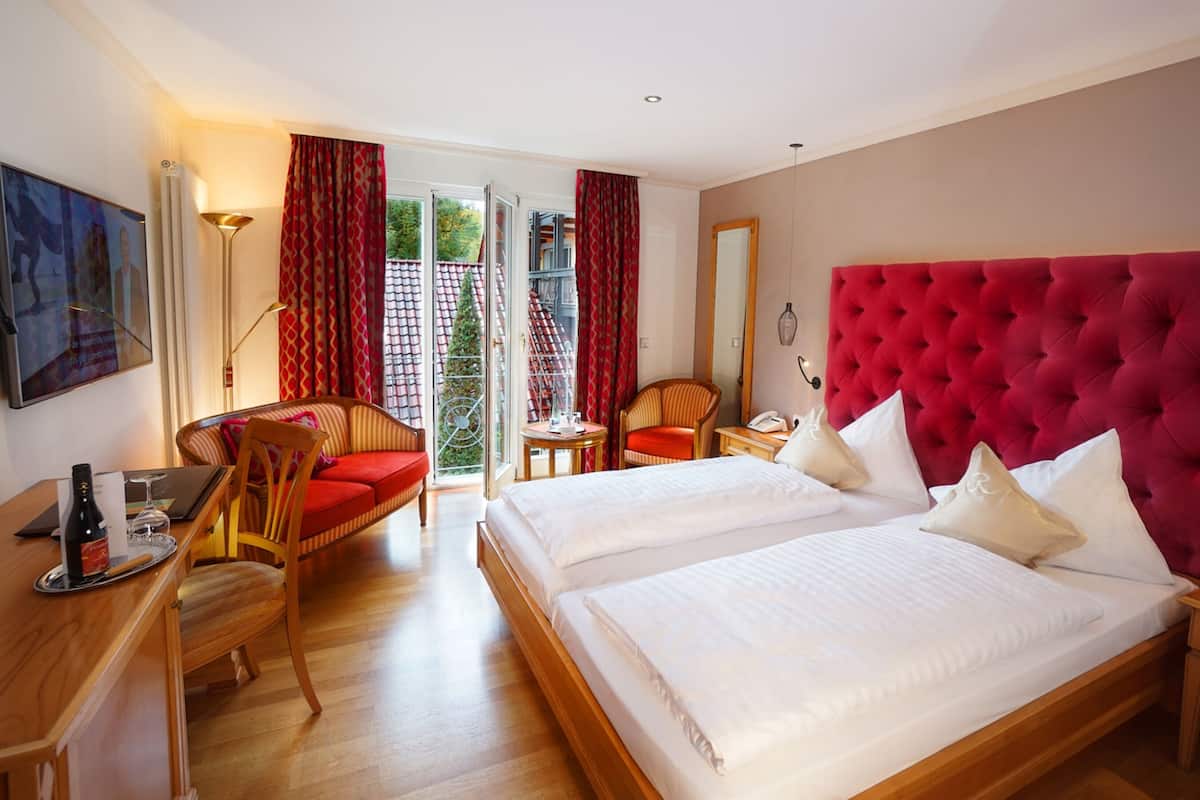 Ein Hotelzimmer mit zwei Betten, einem roten, gepolsterten Kopfteil, einem Holztisch, einem Stuhl, einem kleinen Tisch und einem Balkon mit roten Vorhängen.
