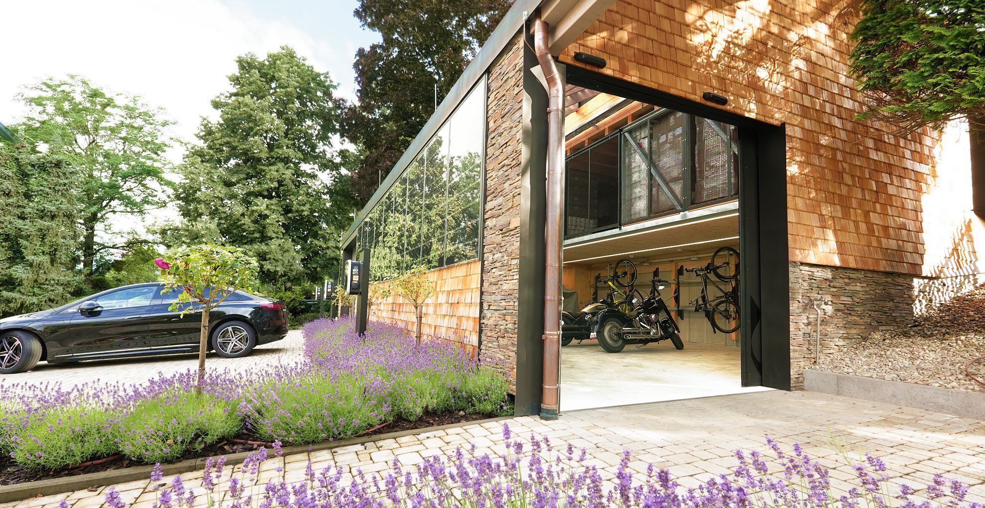 En moderne bygning med garage, en parkeret bil og en række lilla blomster i forgrunden.