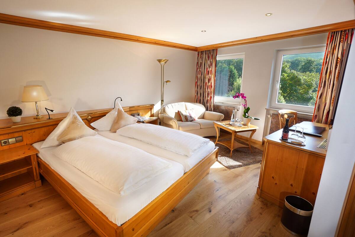 Ein Hotelzimmer mit einem hölzernen Bett, einem Schreibtisch mit Lampe, einem Sessel, einem kleinen Tisch mit Pflanze und einem Fenster mit Vorhängen.