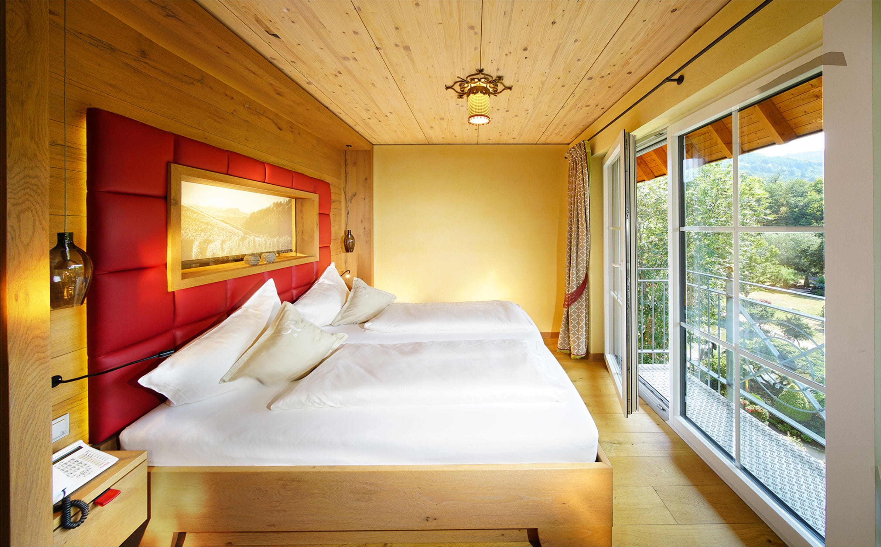Ein Schlafzimmer mit Holzboden, einem großen Bett mit weißer Bettwäsche, einem roten Kopfteil und einem großen Fenster mit Blick auf Grünflächen.