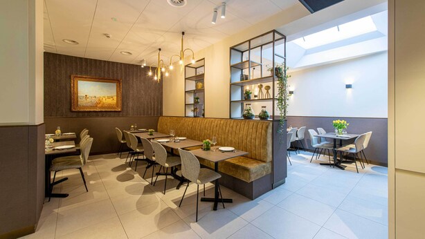 Un espace de salle à manger moderne avec un plafond haut, un éclairage encastré, un coin salon central doté d’un banc rembourré et une étagère murale agrémentée d’objets décoratifs.