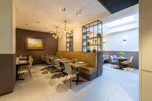 Een moderne eetruimte met een hoge plafond, ingebouwde verlichting, een centraal zithoek met een comfortabele bank, en een wandplank met decoratieve objecten.