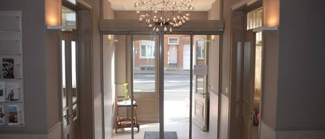 A hotel entrance with a modern chandelier, a glass door leading outside, and a small table with a chair inside.