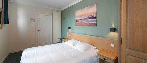 A hotel room with a large bed, a nightstand, a wooden wardrobe, a painting on the wall, and a lamp.