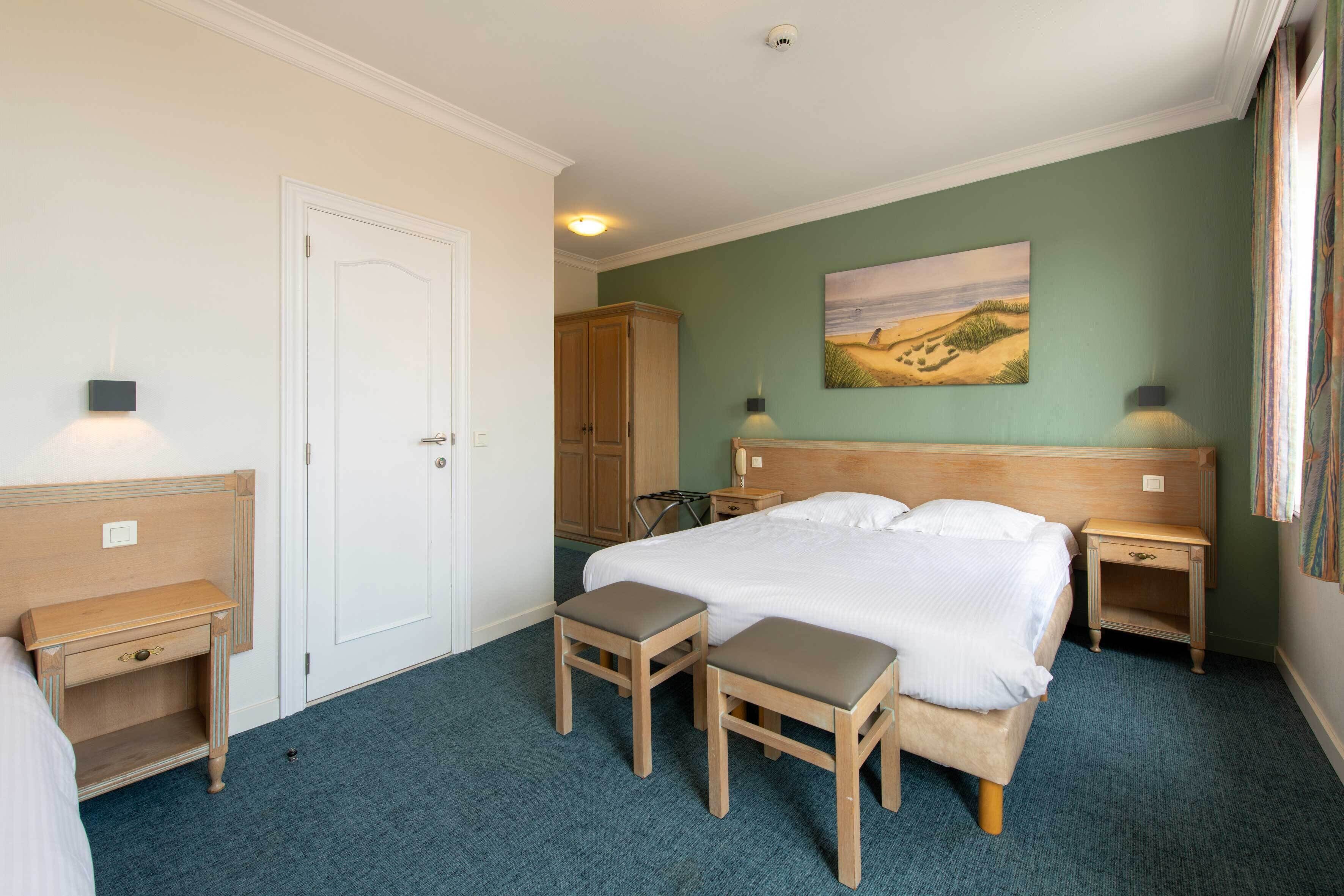 Et hotelværelse med en stor seng, to sengeborde, et lille skrivebord, en stol, en dør og et billede på væggen.