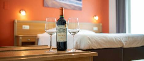 A bottle of Chateau Pascaud wine and two glasses on a wooden table in a hotel room with a bed and a painting on the wall.
