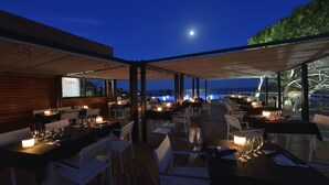 An outdoor dining area with tables set for two, under a covered roof, overlooking a body of water. - Las Colinas Golf & Country Club (Orihuela)
