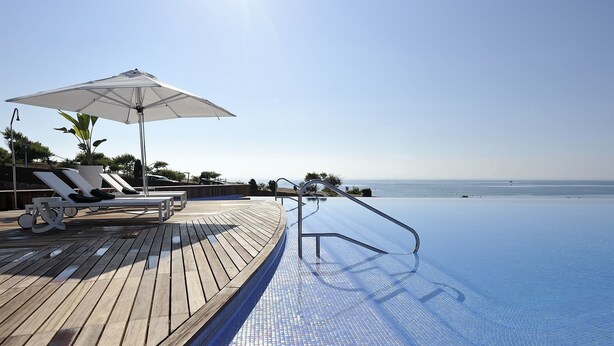 An infinity pool with a wooden deck, lounge chairs, and a large umbrella. - Las Colinas Golf & Country Club (Orihuela)
