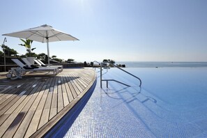 Una piscina a sfioro con un solarium in legno, sedie a sdraio e un ombrellone grande.