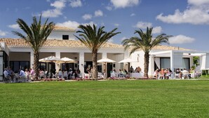 An outdoor dining area with tables and chairs, palm trees, and a building with a tiled roof. - Las Colinas Golf & Country Club (Orihuela)