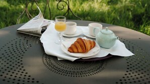 Et bord med en croissant, en tepotte og et glas appelsinjuice, placeret udendørs ved et metalbord med et hus i baggrunden.