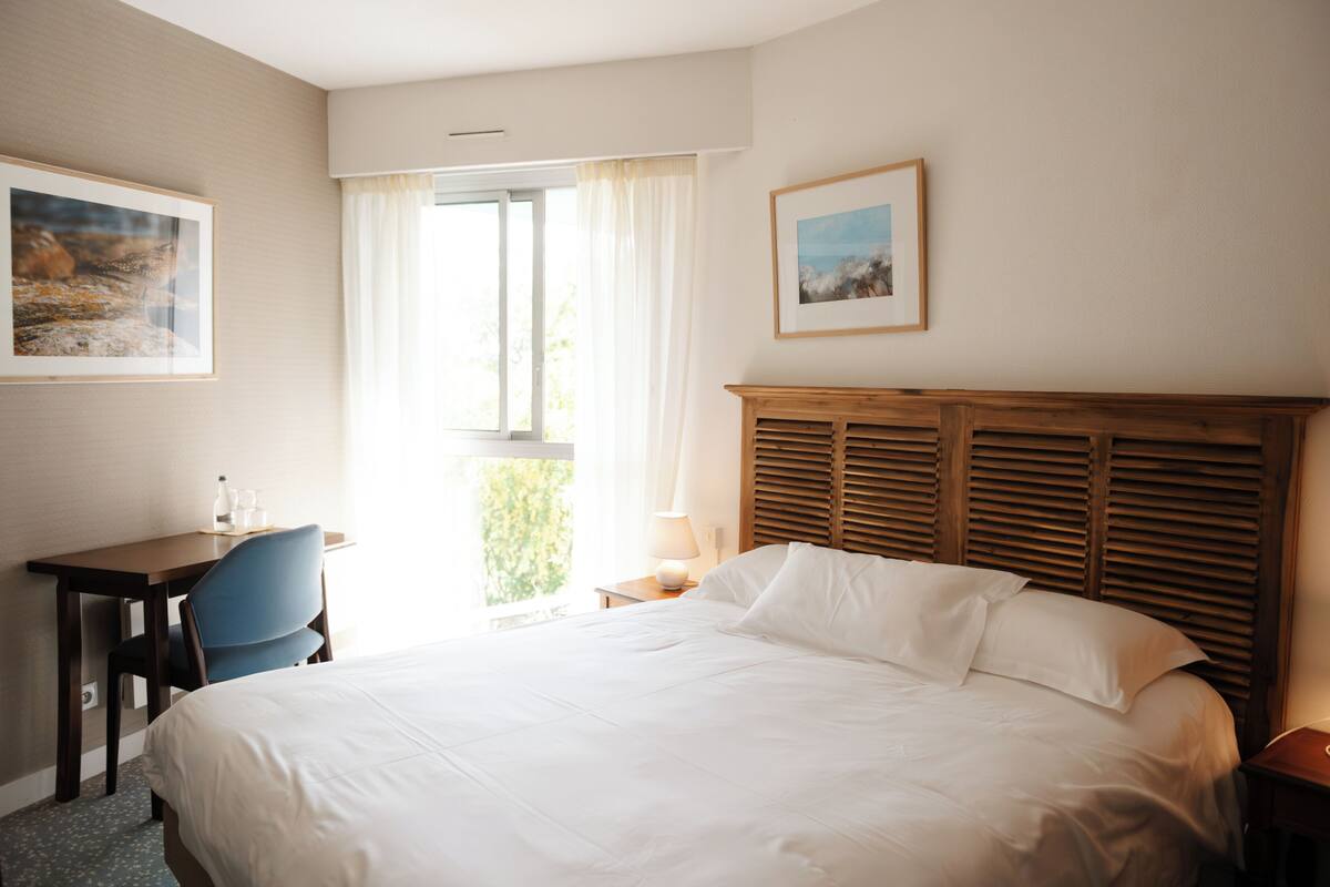A hotel room with a bed, a desk, a chair, a window with curtains, and two framed pictures on the wall.