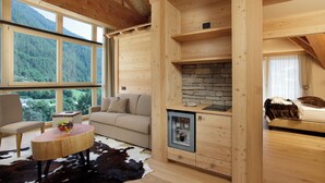 A cozy wooden cabin interior with a stone fireplace, a round wooden table with fruit, a sofa, and a bed with a cat.