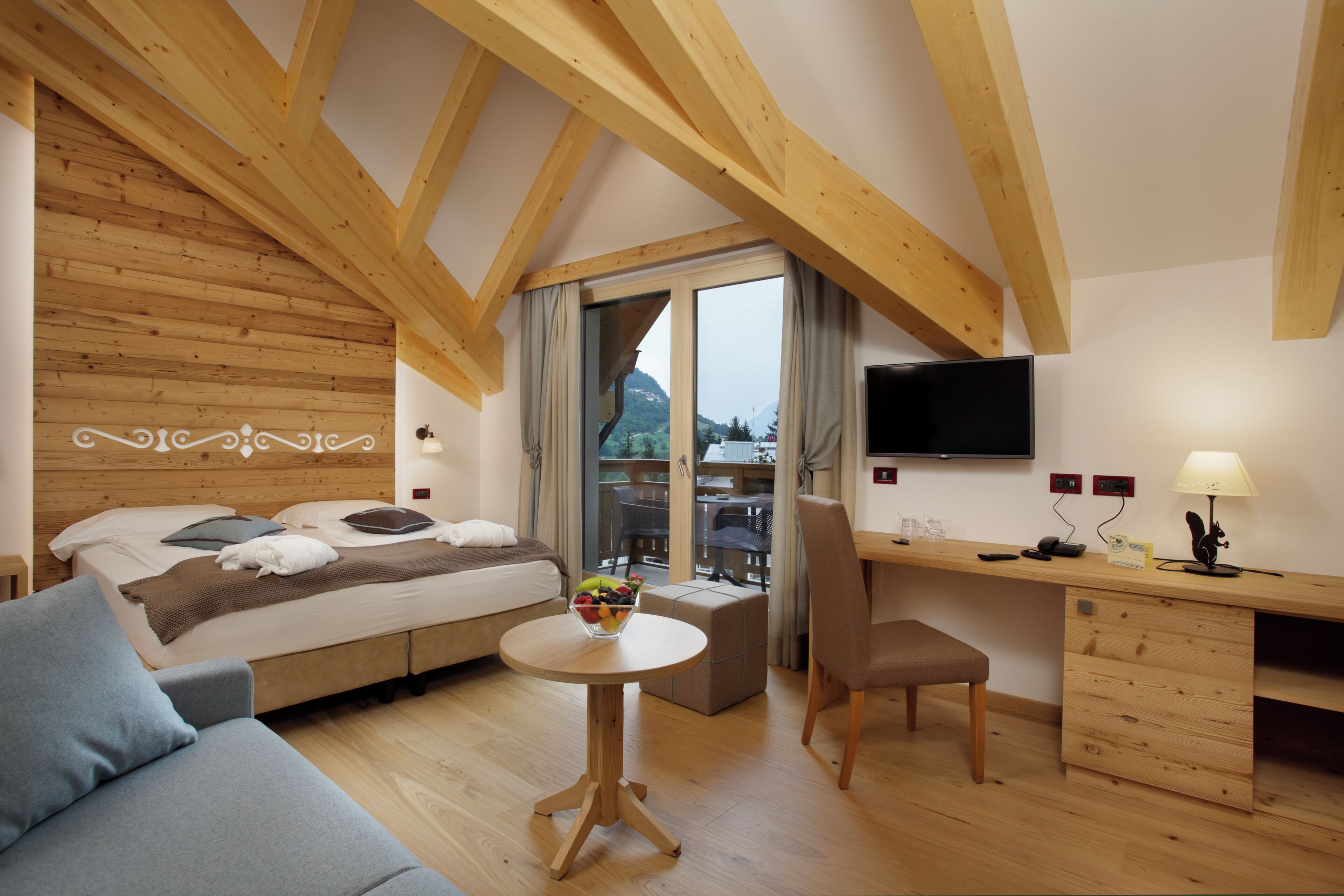 Ein modernes Schlafzimmer mit Holzbalkendecke, einem Bett mit Kissen, einem Nachttisch mit Obstschale, einem Schreibtisch mit Lampe und einem Fernseher an der Wand.