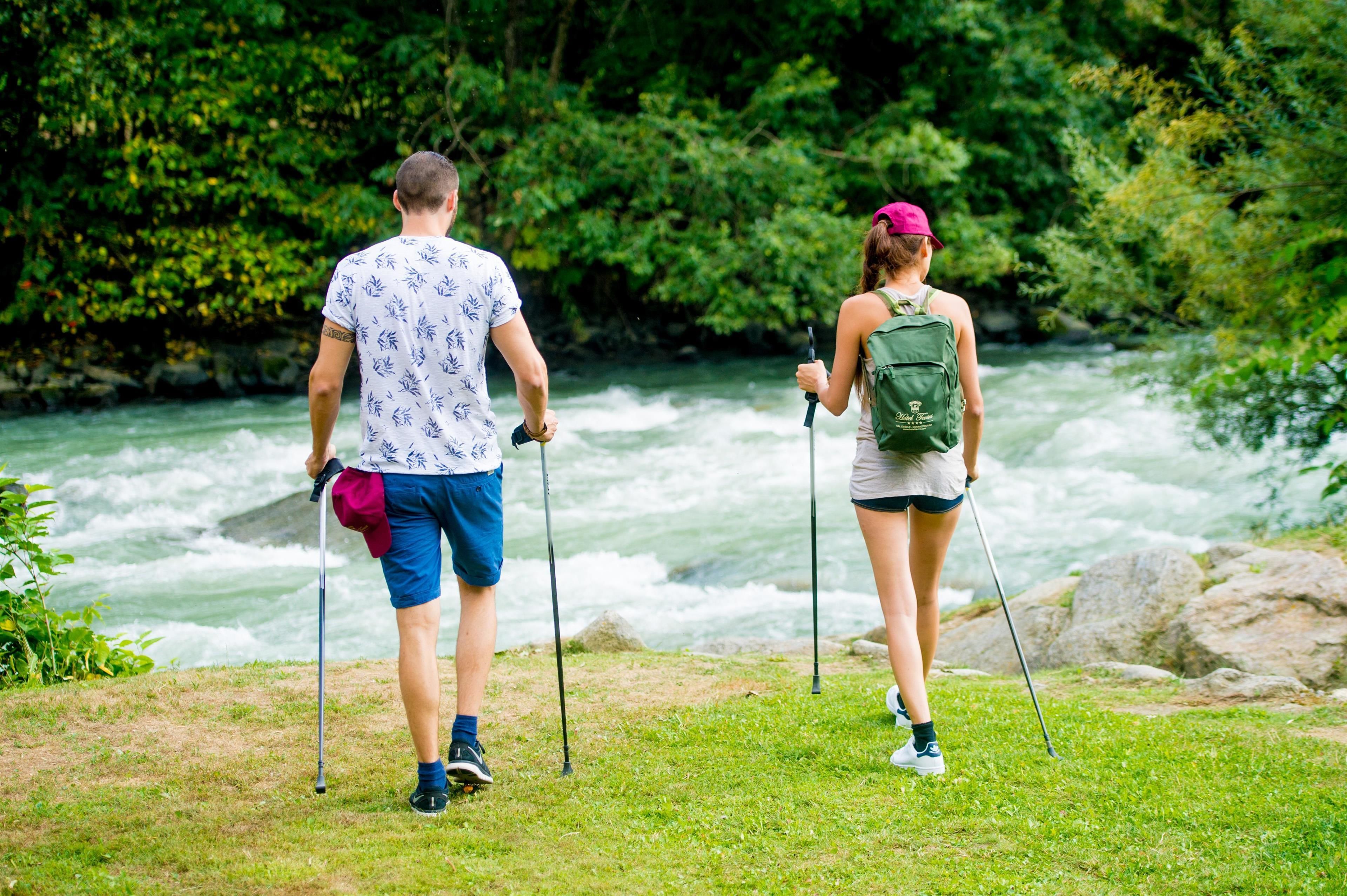 Dos personas caminando con bastones de trekking cerca de un río, rodeadas de vegetación.