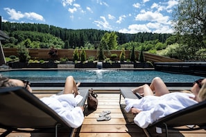 Zona junto a la piscina con sillones de descanso y una terraza de madera con vistas a una piscina.