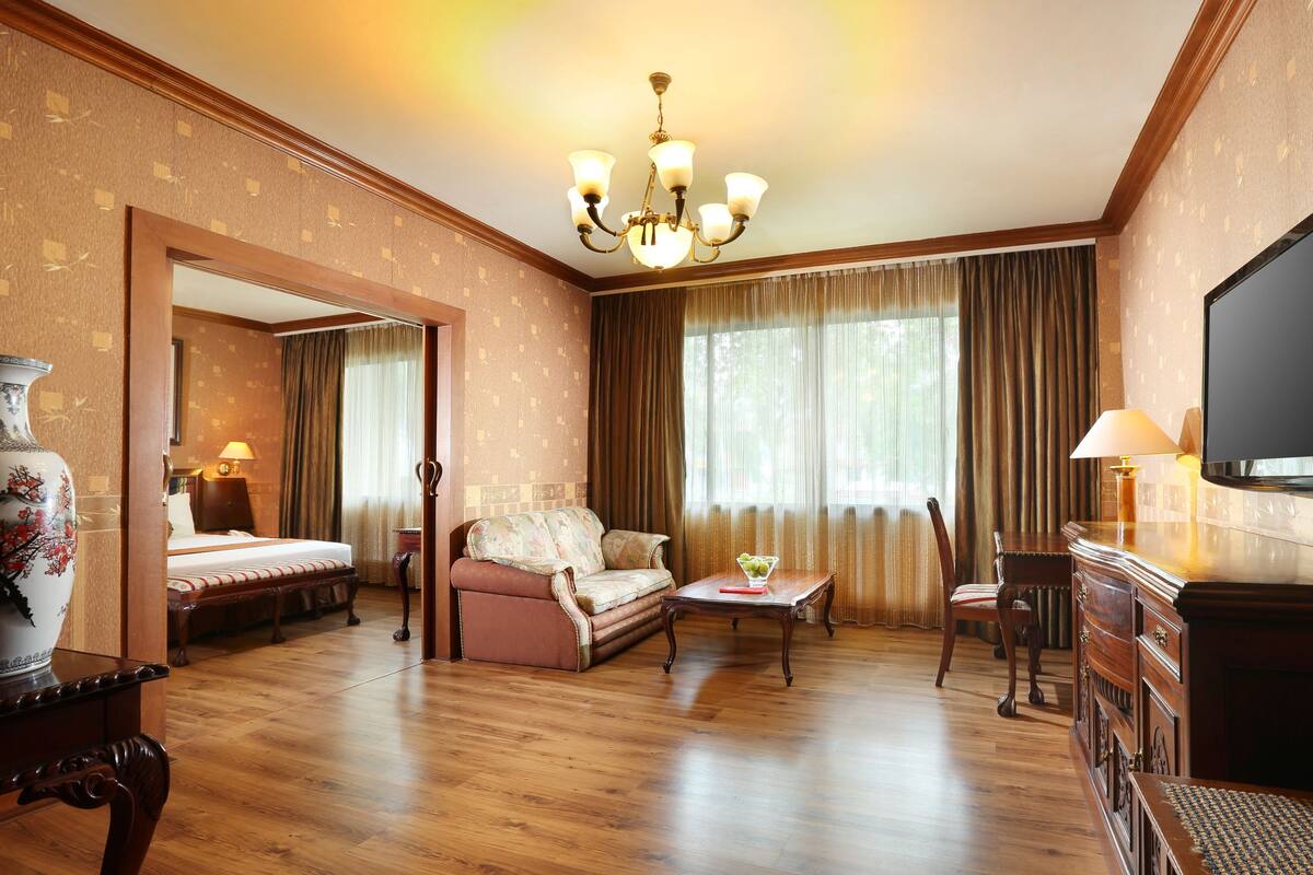 Habitación de hotel con piso de madera, un sofá, una silla, un televisor, una lámpara y un florero.
