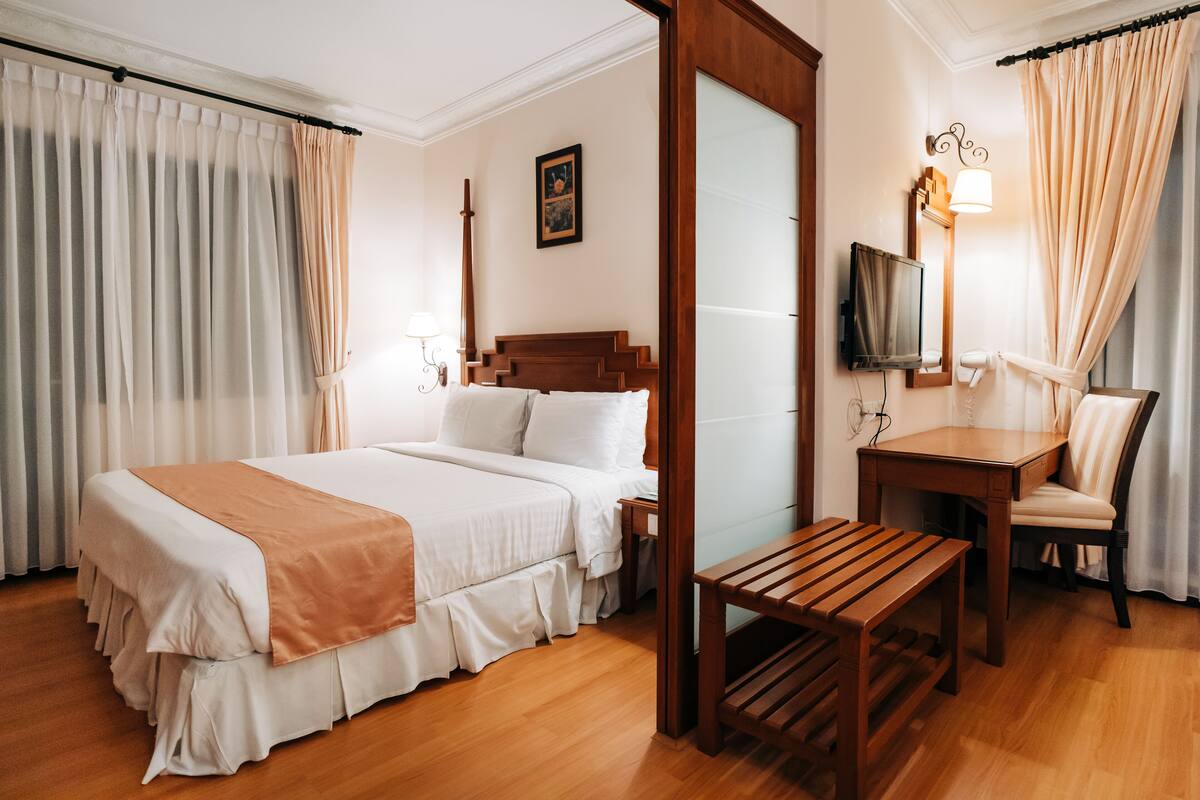 A hotel room with a large bed, a wooden bench, a desk with a chair, a television, and a mirror.
