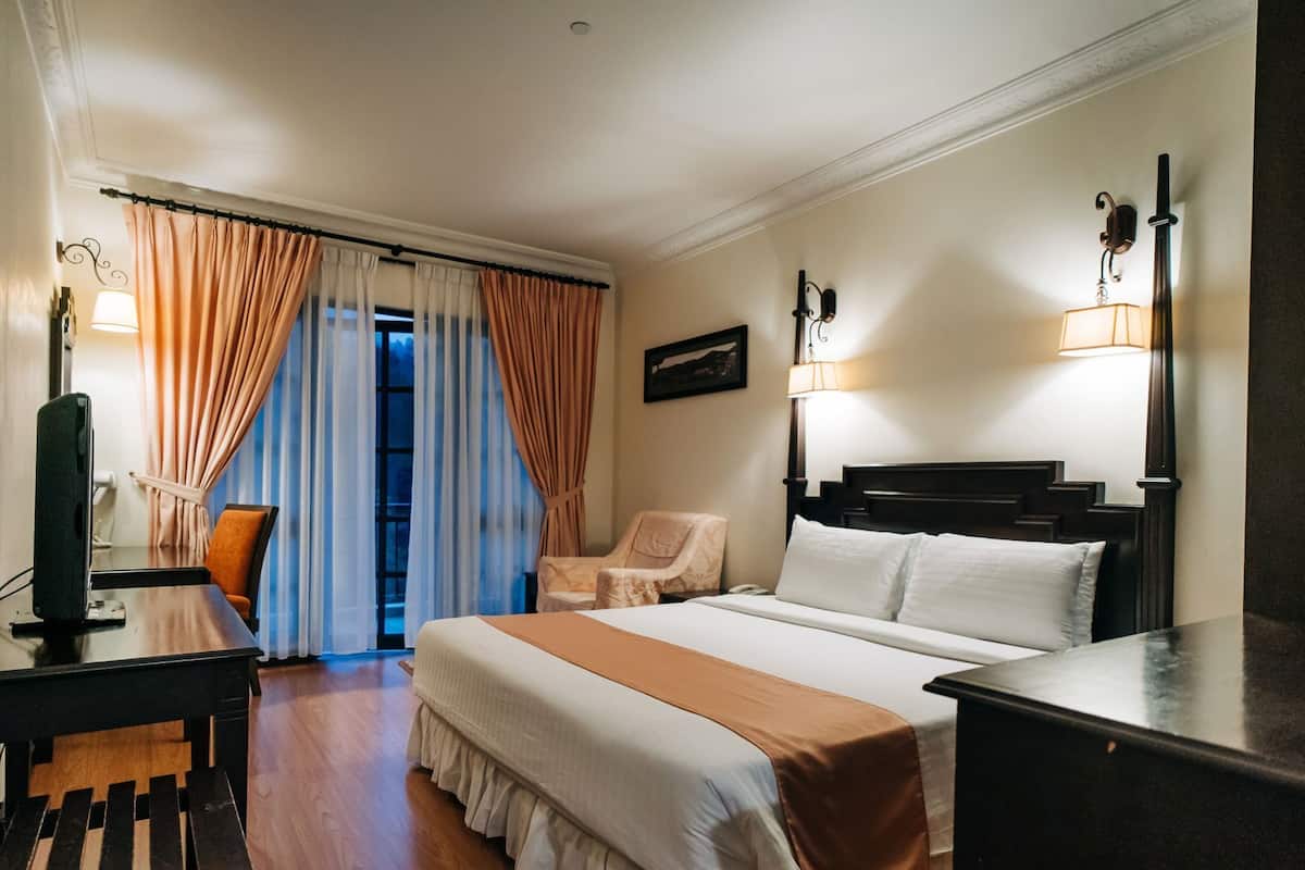 A hotel room with a bed, a desk with a computer, a chair, a lamp, and a window with curtains.