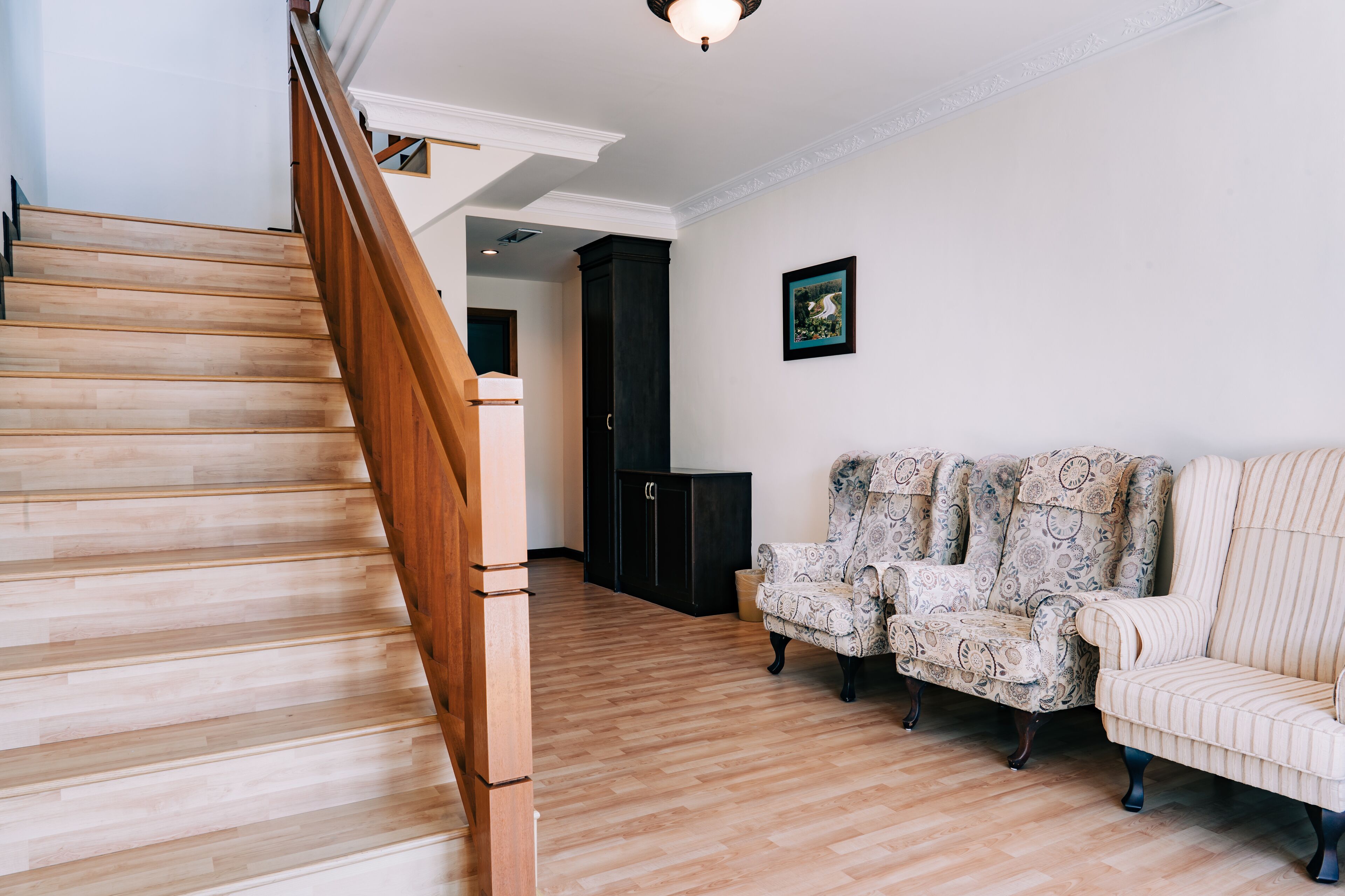 A staircase with wooden steps and a handrail, leading to a dark-colored room with patterned armchairs and a framed picture on the wall.