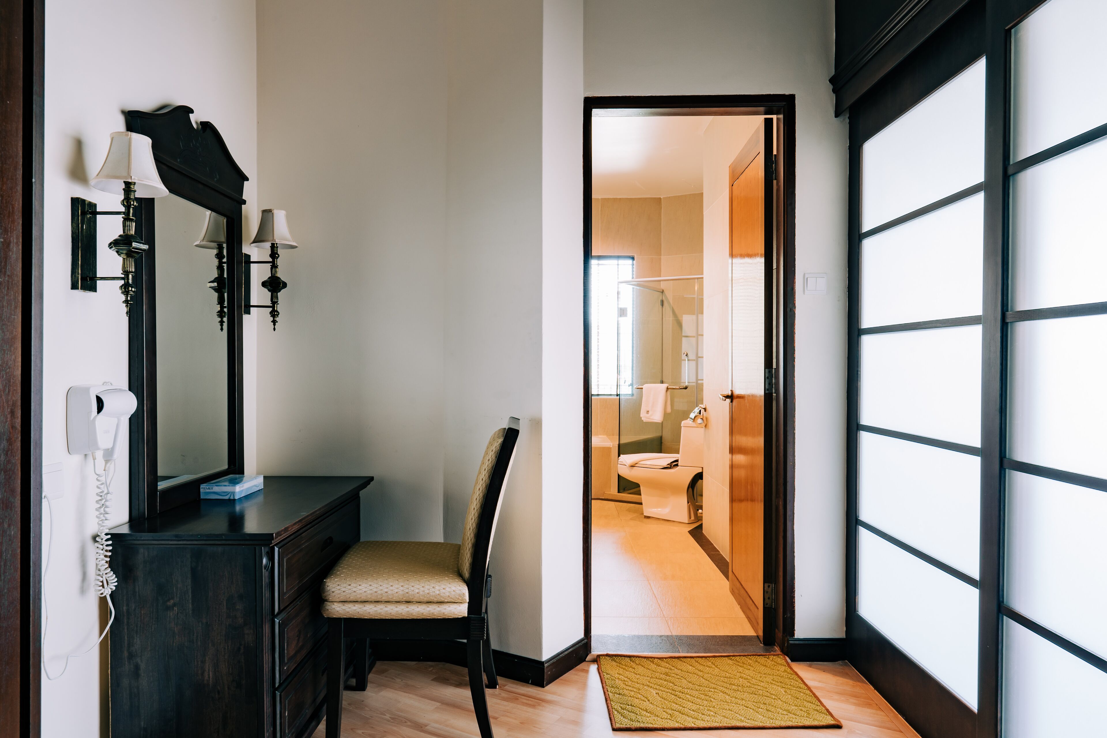 Habitación de hotel con piso de madera, cómoda de madera oscura con espejo y lámparas de pared, una silla y vista al baño a través de una puerta abierta.
