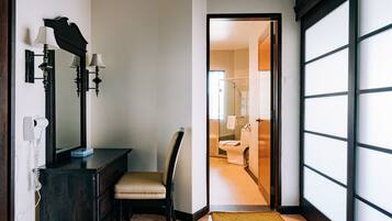 Habitación de hotel con piso de madera, cómoda de madera oscura con espejo y lámparas de pared, una silla y vista al baño a través de una puerta abierta.