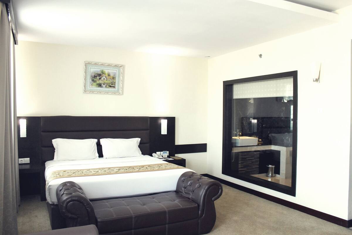 A hotel room with a large bed, a bedside table, a nightstand, a small sofa, a large mirror, and a picture on the wall.